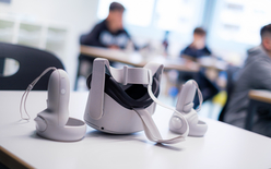 Im Vordergrund eine VR-Brille mit den Handsticks liegen auf dem Tisch, im Hintergrund sind Kinder erkennbar.